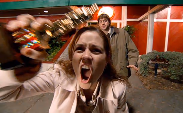 Pam Beesly yelling and holding up a Dundie Award trophy while Jim Halpert stands behind her outside | Timestripe